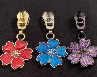Può includere: Tre tiretti per cerniera con ciondoli a forma di fiore. I ciondoli sono rossi, blu e viola con glitter. I tiretti per cerniera sono dorati, argentati e neri.