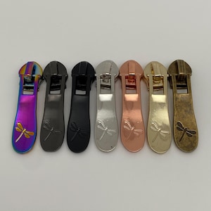 Könnte beinhalten: Sieben Reißverschluss-Zipper mit Libellen-Design. Die Zipper sind in verschiedenen Ausführungen erhältlich, darunter Regenbogen, Schwarz, Silber, Kupfer, Gold und Antikmessing.