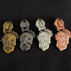 Könnte beinhalten: Vier Reißverschluss-Zipper mit Totenkopf- und Blumendesign. Die Reißverschluss-Zipper sind in Gold, Silber und Roségold erhältlich.
