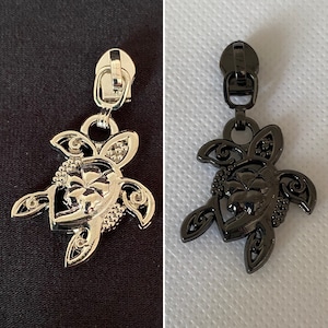 Könnte beinhalten: Zwei Reißverschluss-Zipper in Form einer Schildkröte mit Blumendesign. Einer ist silber und der andere ist schwarz.