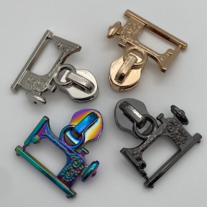 Könnte beinhalten: Vier Metall-Reißverschlussanhänger in Silber, Gold, schillernd und Schwarz. Jeder Anhänger hat ein dekoratives Design, wobei die schillernden und schwarzen Anhänger wie Nähmaschinen geformt sind. Die Anhänger haben eine Schlaufe zum Befestigen an einem Reißverschluss.