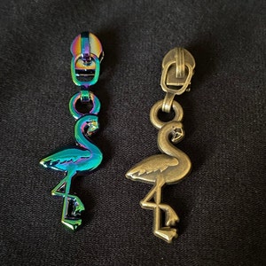Könnte beinhalten: Zwei Metall-Reißverschluss-Zipper in Form von Flamingos. Einer ist regenbogenfarben und der andere ist antikes Gold.