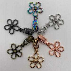 Könnte beinhalten: Sechs verschiedene Reißverschluss-Zipper mit blütenförmigen Anhängern. Die Anhänger sind in Silber, Gold und Roségold. Die Reißverschluss-Zipper sind in Silber, Gold und Roségold.