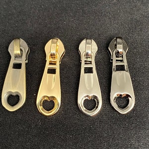 Könnte beinhalten: Vier Reißverschluss-Zipper mit herzförmigen Ausschnitten in den Ausführungen Antikmessing, Gold, Silber und Schwarz.