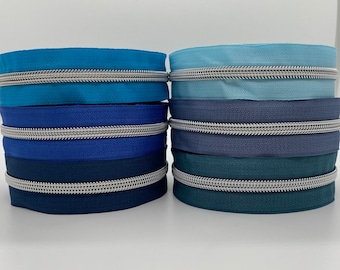 May include: A stack of blue, teal, and gray zipper tape with silver teeth. The tape is arranged in a gradient from light blue to dark gray.