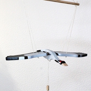 Peut inclure: Un mobile en bois en forme de grue avec des ailes noires et blanches, un corps gris et un bec rouge. La grue est suspendue à une baguette en bois par des cordes et a un poids en liège suspendu à son ventre.