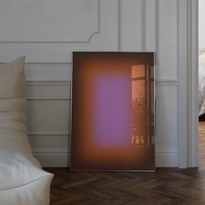 Könnte beinhalten: Ein gerahmter abstrakter Kunstdruck mit Farbverlauf. Das Kunstwerk zeigt eine Mischung aus Orange-, Lila- und Brauntönen. Der Druck lehnt an einer weißen Wand mit Leisten, und der Boden ist aus dunklem Holz.