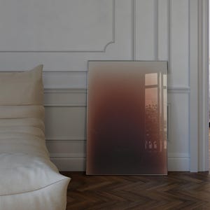 May include: A rectangular framed artwork with a colour gradient, shifting from dark brown to reddish-brown and beige. It leans against a white wall in a room with a bed and a window, with a wooden floor.