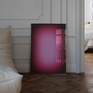May include: A framed art print with a pink and purple gradient. The artwork is on a wooden floor, propped against a white wall with moulding. A bedroom with a bed is visible in the background, with a glimpse of a window.