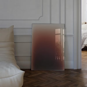May include: A large, rectangular framed artwork with a gradient of colours from dark brown to light brown. The artwork is leaning against a white wall with moulding, and the floor is a dark wood. The artwork reflects the light from a window.
