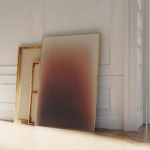 May include: Three framed canvases lean against a white wall. The largest canvas features a gradient of colours, transitioning from light to dark. The frames have a gold finish. The room has a hardwood floor and a white door.