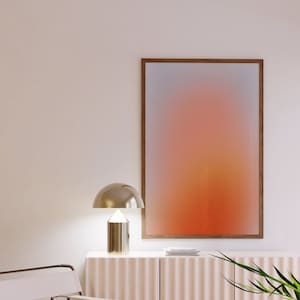 Könnte beinhalten: Ein weißer Stuhl mit einem Metallgestell steht vor einem weißen Schrank mit einem Holzrahmen-Abstraktbild darüber. Der Druck zeigt einen Farbverlauf von Orange, Gelb und Rosa. Eine goldene Tischlampe mit einem runden Schirm steht auf dem Schrank.