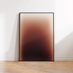 May include: A framed art print with an abstract design. The colours blend from a light beige at the top to a deep burgundy at the bottom. The artwork is displayed against a white wall, leaning on a wooden floor.