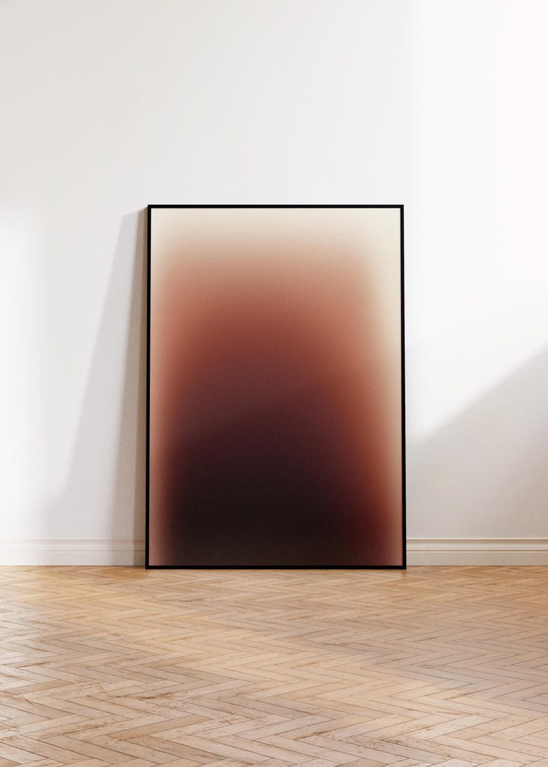 May include: A framed art print with an abstract design. The colours blend from a light beige at the top to a deep burgundy at the bottom. The artwork is displayed against a white wall, leaning on a wooden floor.
