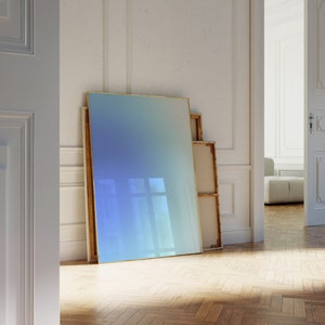 May include: A large, framed, blue and white gradient painting leans against a white wall in a room with wood floors. The painting is framed in a thin, gold frame.