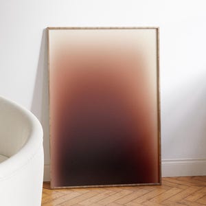 May include: A framed artwork with a gradient design. The colour transitions from a light beige at the top to a deep burgundy at the bottom. The frame is a light brown colour. The artwork is leaning against a white wall.