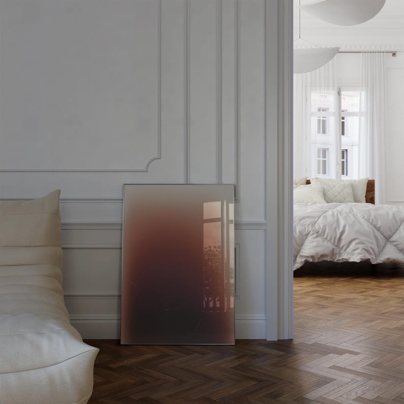 May include: A rectangular framed artwork with a colour gradient, shifting from dark brown to reddish-brown and beige. It leans against a white wall in a room with a bed and a window, with a wooden floor.