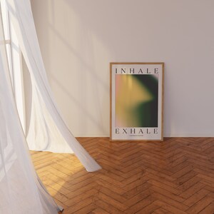 May include: A framed poster with the words "INHALE" and "EXHALE" in a minimalist font. The artwork features a blurred gradient of pink, yellow, and green. The poster is displayed on a wooden floor, next to a sheer white curtain.