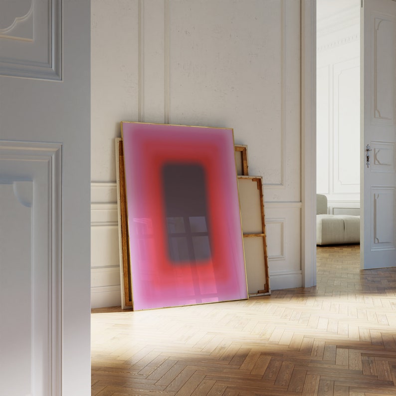 May include: A framed abstract art print with a gradient of pink and red colours. The print has a thin gold frame and is leaning against a white wall.