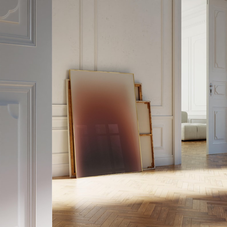 May include: A large, rectangular artwork with a gradient of colours from light beige to deep brown, leaning against a white wall. The artwork has a thin gold frame. Other framed canvases are stacked behind it. The room has a hardwood floor.