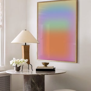 May include: A round marble table with a wooden base and a lamp with a white shade. There are two white stools and a framed abstract art print with a gradient of pink, orange, yellow, green, and blue colours. The table has a stack of books and a small gold bowl on top.