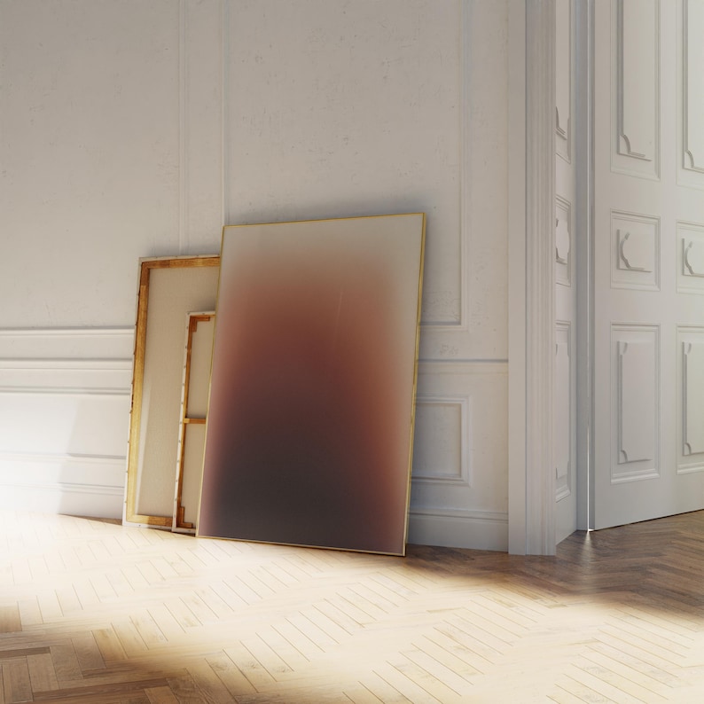 May include: Three framed canvases lean against a white wall. The largest canvas features a gradient of colours, transitioning from light to dark. The frames have a gold finish. The room has a hardwood floor and a white door.