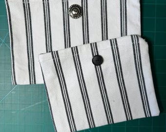 Peut inclure: Deux pochettes rectangulaires en tissu à fond blanc avec des rayures verticales noires. La plus grande pochette est dotée d'une fermeture à bouton argenté métallique, tandis que la plus petite possède un bouton noir simple.