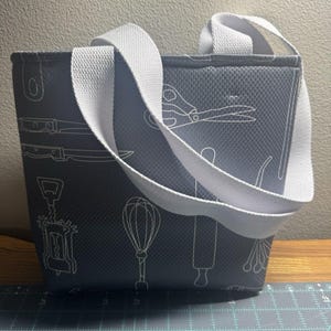 May include: A gray tote bag with a white diamond-textured pattern and white line art illustrations of kitchen tools including scissors, a whisk, a rolling pin, and a corkscrew. The bag features two wide white fabric handles attached at the top.