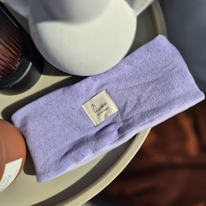Könnte beinhalten: Ein lavendelfarbenes Stirnband mit einem kleinen rechteckigen Etikett mit der Aufschrift "Le gentil by Julia Steger". Das Stirnband ist aus einem weichen, strukturierten Stoff gefertigt und liegt auf einem hellen Tablett.