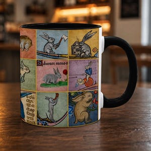 May include: A white ceramic mug with a black handle and interior, featuring a grid of colourful medieval-style illustrations of rabbits in various activities, including one holding an axe, one with a shield, and text in a gothic script.