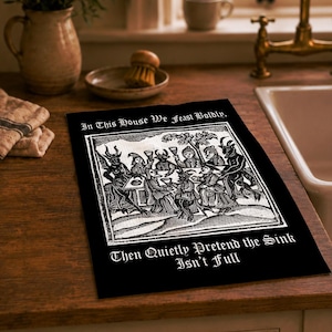Könnte beinhalten: Ein rechteckiges schwarzes Küchentuch mit einer weißen Illustration im Holzschnitt-Stil, die Figuren bei einem Festmahl zeigt. Der Text lautet: In This House We Feast Boldly, Then Quietly Pretend the Sink Isn't Full. Das Motiv ist mittig platziert.