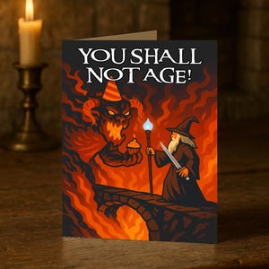 May include: Greeting card with the text "YOU SHALL NOT AGE!" depicts a wizard and a demon in a fiery scene. The wizard holds a staff with a blue flame and a sword. The demon holds a cupcake.