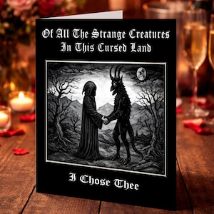 May include: A black greeting card with the text "Of All The Strange Creatures In This Cursed Land I Chose Thee." The card features a black and white illustration of two figures shaking hands in a dark forest setting. The background includes candles, flowers, and champagne glasses.