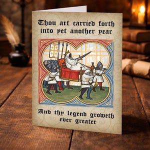 Op de afbeelding: Wenskaart met middeleeuwse illustratie en tekst. De kaart toont figuren die een persoon in een gewaad dragen, met de zinnen "Thou art carried forth into yet another year" en "And thy legend groweth ever greater."