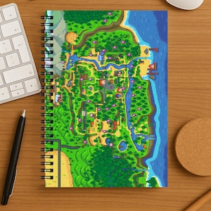 May include: Spiral-bound notebook with a pixel art map of a rural landscape, including rivers, forests, and buildings. The map uses green, blue, and brown. The notebook is on a wooden desk with a keyboard, glasses, and a pen. The measurements are in cm.