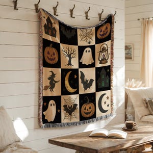 May include: A Halloween-themed throw blanket with a patchwork design. The squares feature spooky illustrations of pumpkins, bats, ghosts, and haunted houses in black, tan, and orange. The blanket is hanging on a white wall with metal hooks.