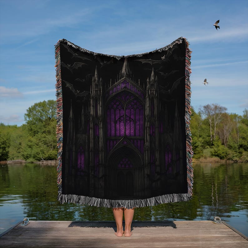 Puede incluir: Manta negra con un dise&ntilde;o de catedral g&oacute;tica, vidrieras moradas y siluetas de murci&eacute;lagos. La manta tiene un borde con flecos. El fondo muestra un lago y un cielo azul.