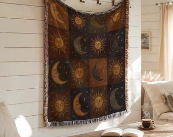 May include: A woven tapestry blanket featuring a celestial design of suns and moons in a patchwork pattern. The blanket has a fringed edge and is displayed on a white wall. The color palette includes shades of brown, blue, and gold.