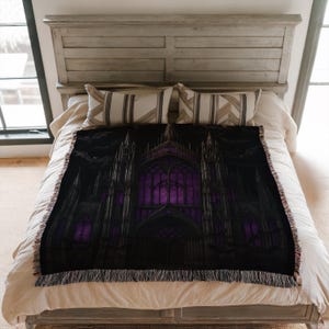 Puede incluir: Una manta negra con un dise&ntilde;o de catedral g&oacute;tica y vidrieras moradas. La manta tiene un borde con flecos y se muestra en una cama con ropa de cama de tonos neutros.