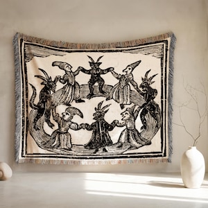 Puede incluir: Un tapiz tejido con una ilustración en blanco y negro de figuras con cuernos y sombreros puntiagudos bailando en círculo. El tapiz tiene un fondo crema, un borde negro y un borde con flecos.