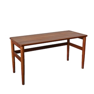 Puede incluir: Una mesa de centro de madera de teca de estilo moderno de mediados de siglo con una tapa rectangular y un solo travesaño debajo. La mesa es de color marrón cálido y tiene un diseño sencillo.