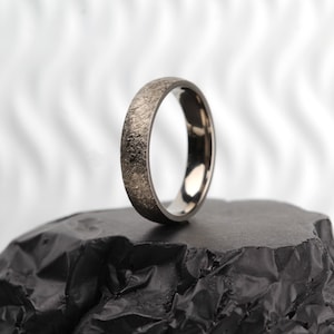 May include: A silver ring with a textured, brushed finish. The ring is sitting on a black, textured surface.