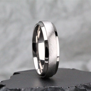 Op de afbeelding: Een zilverkleurige ring met een geborstelde middenband en gepolijste randen. De ring heeft een eenvoudig, modern ontwerp, geschikt voor dagelijks gebruik. De ring wordt getoond op een donkere ondergrond.