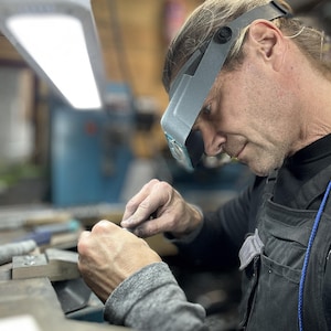 May include: A person wearing a grey head-mounted magnifying visor with integrated lights while working on a small object at a workbench. The individual is dressed in a black shirt and a dark grey apron, focusing intently on the task at hand.