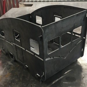 Puede incluir: Un hogar de metal negro con forma de caravana. El hogar tiene una base rectangular y un techo curvo. Hay recortes para ventanas y una puerta en el lateral.