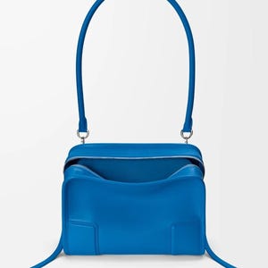 Pode incluir: Uma mala azul vibrante com uma al&ccedil;a superior curva e uma al&ccedil;a longa e fina. A mala tem formato retangular com fecho de z&iacute;per e design minimalista. A mala &eacute; feita de um material liso.