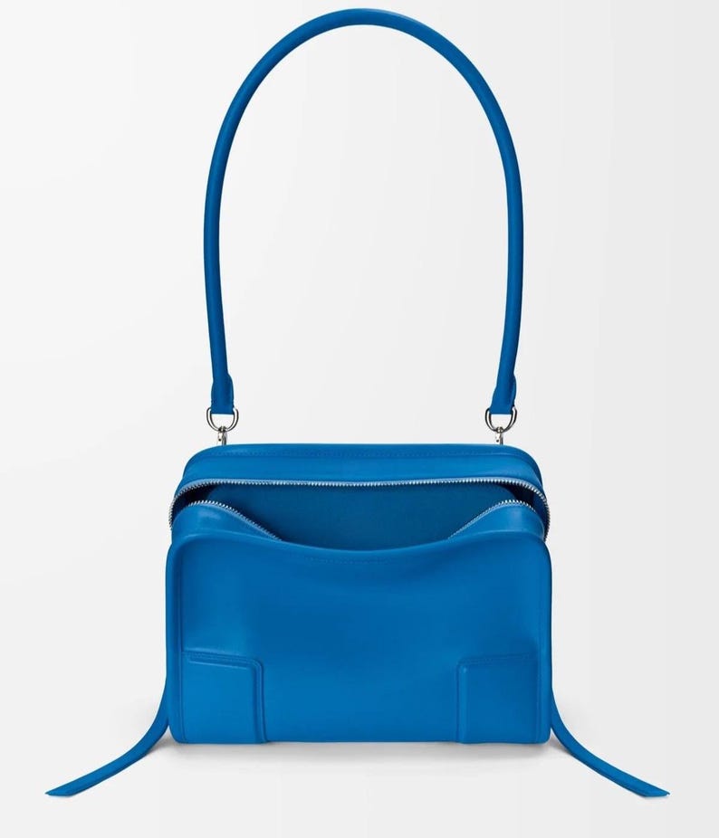 Pode incluir: Uma mala azul vibrante com uma al&ccedil;a superior curva e uma al&ccedil;a longa e fina. A mala tem formato retangular com fecho de z&iacute;per e design minimalista. A mala &eacute; feita de um material liso.