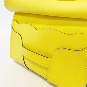 May include: A bright yellow leather bag featuring a smooth texture and white contrast stitching. The front flap displays a distinct scalloped edge design with multiple layered pockets, showcasing clean lines and a structured, minimalist construction.