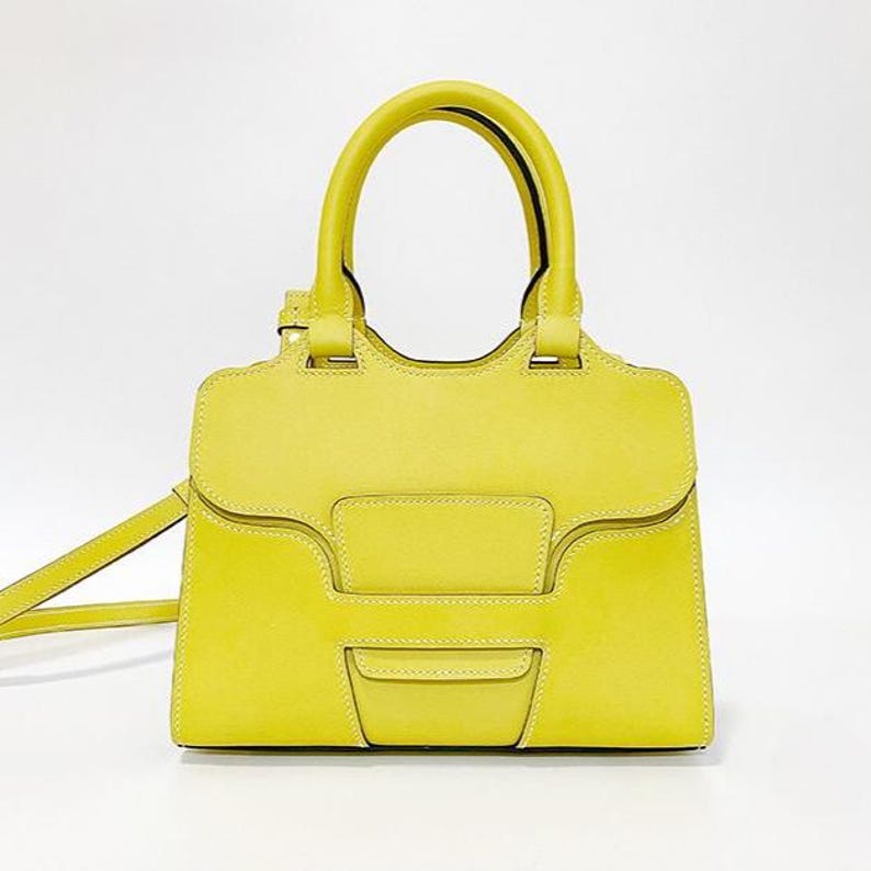 May include: A bright yellow structured handbag with a smooth leather-like finish, featuring two top handles, a flap closure with a central geometric panel, visible white stitching, and a thin detachable shoulder strap extending to the left side.