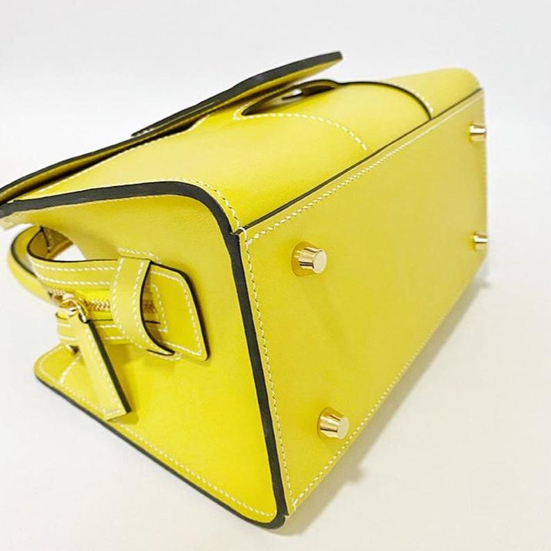 May include: A bright yellow handbag featuring white contrast stitching, black edge paint, and four gold-toned metal feet on the bottom. The side of the bag displays a gold-toned zipper closure with a matching yellow leather pull tab and a strap detail.